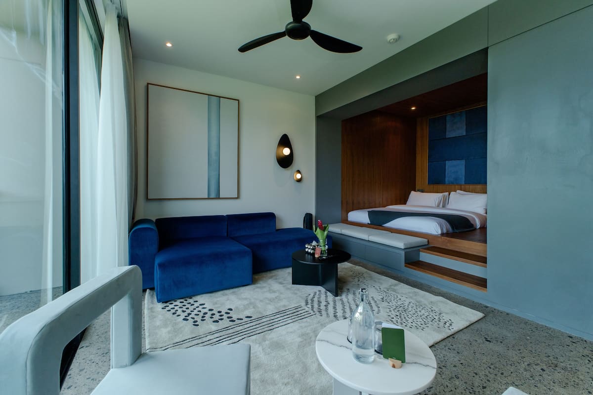 Une chambre d’hôtel moderne avec un canapé bleu, une table ronde blanche, un ventilateur de plafond et un lit doté d’une tête de lit en bois.