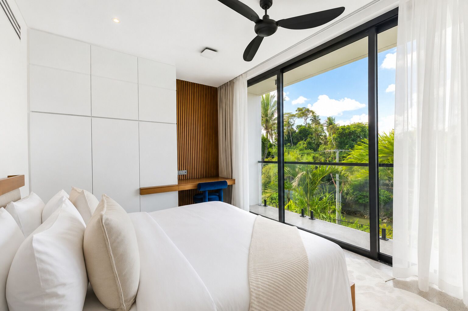 A bedroom with a large bed, a ceiling fan, a view of greenery through a balcony, and a wooden desk.