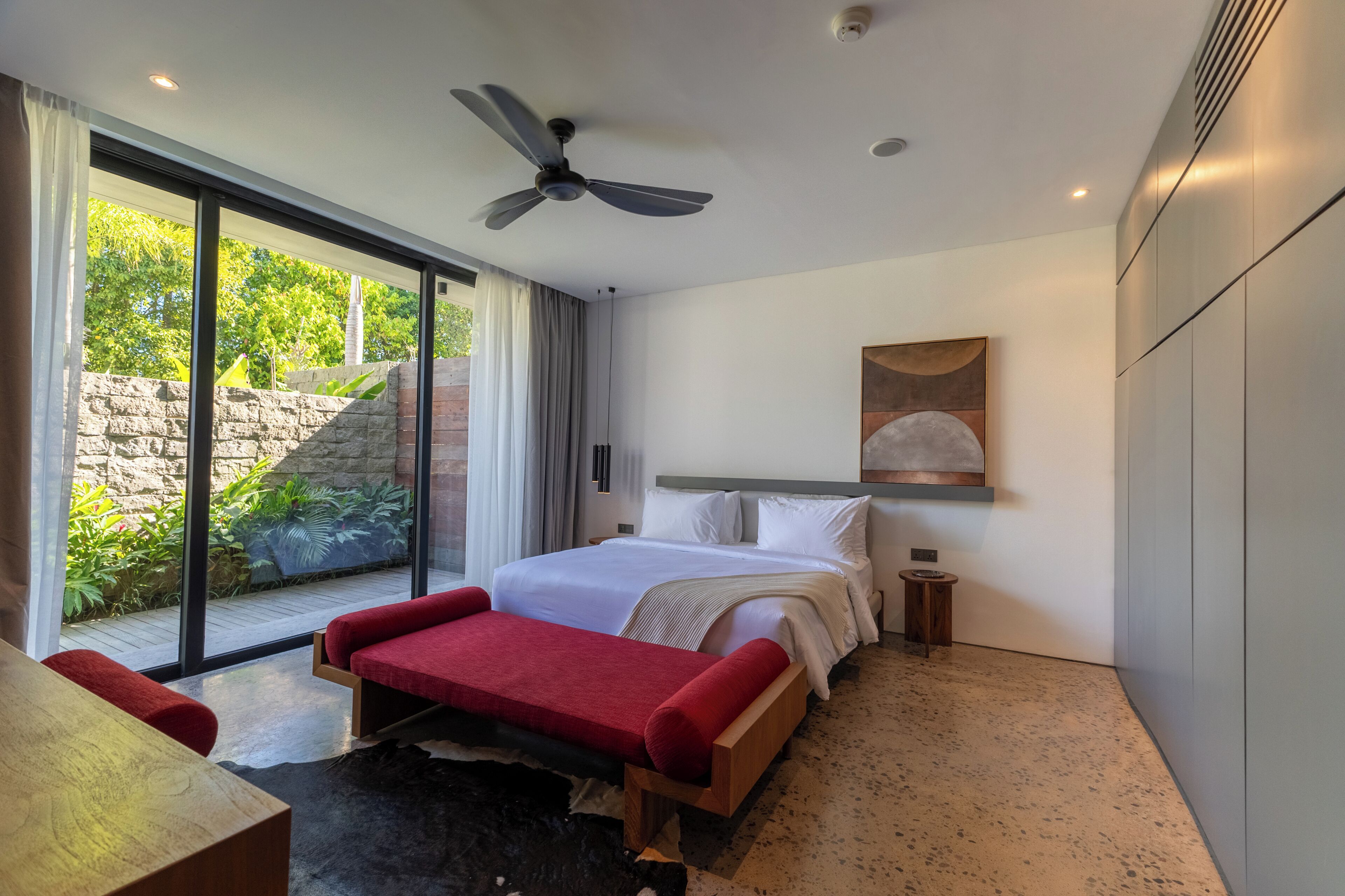 Quarto moderno com cama grande, ventoinha no teto, mesinha de cabeceira e vista para o exterior.