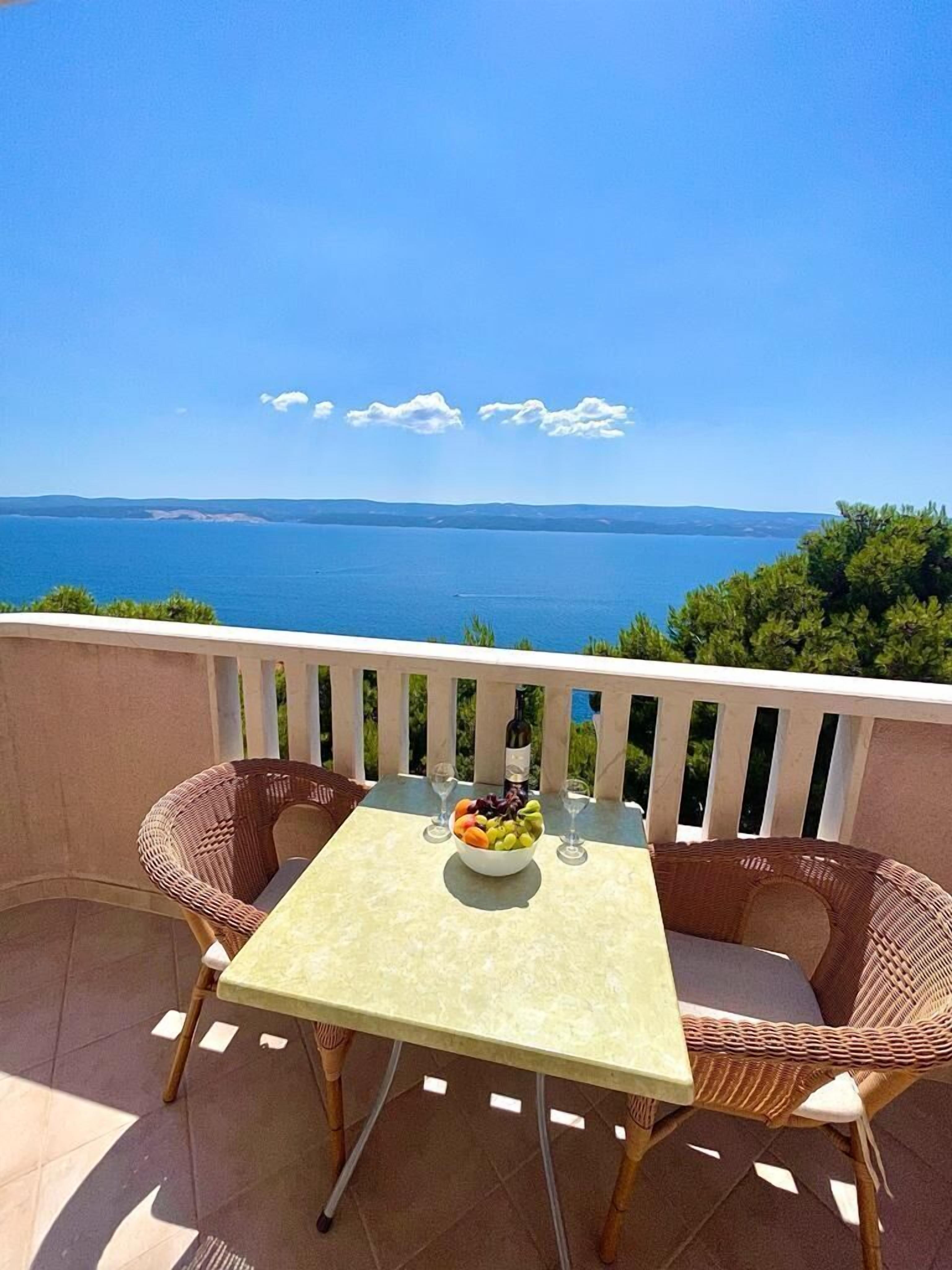 A balcony with a table, two wicker chairs, a bowl of fruit, and a bottle of wine.
