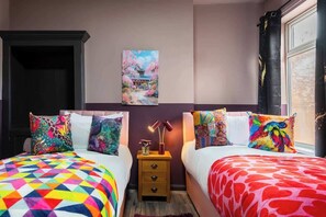 Un dormitorio con dos camas, ropa de cama colorida, una mesita de noche con lámpara y un cuadro en la pared.