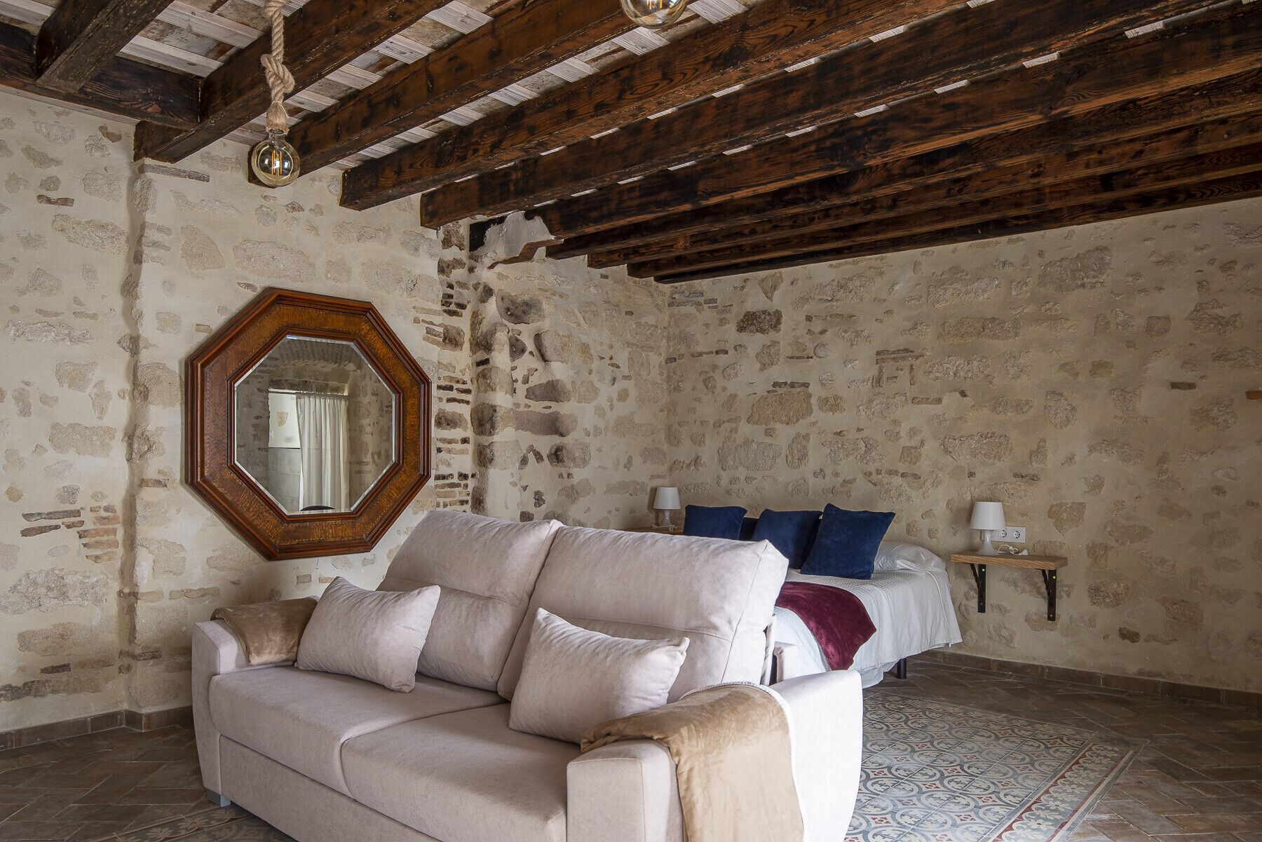 A room with a stone wall, wooden beams, a large mirror, a sofa, a bed with blue pillows, and a small bedside table with a lamp.