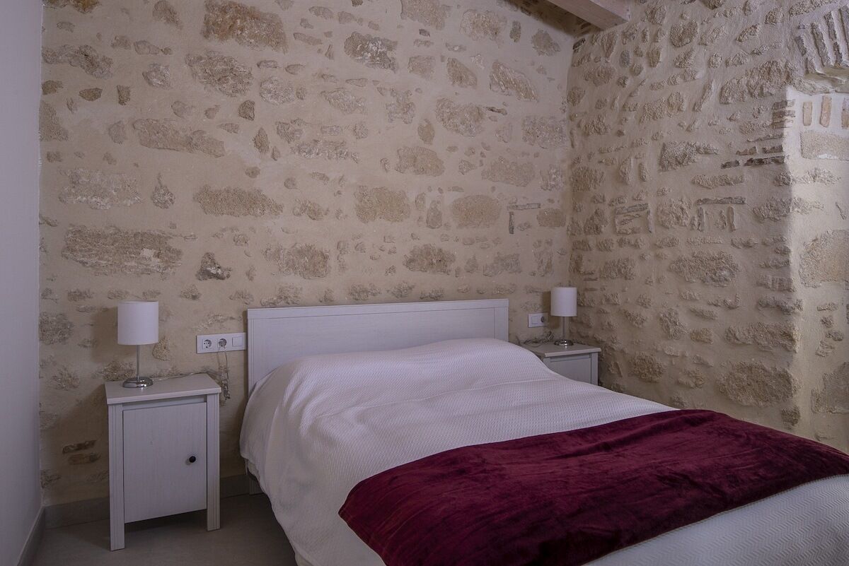 A bedroom with a stone wall, a white bed, a red blanket, and two bedside tables with lamps.