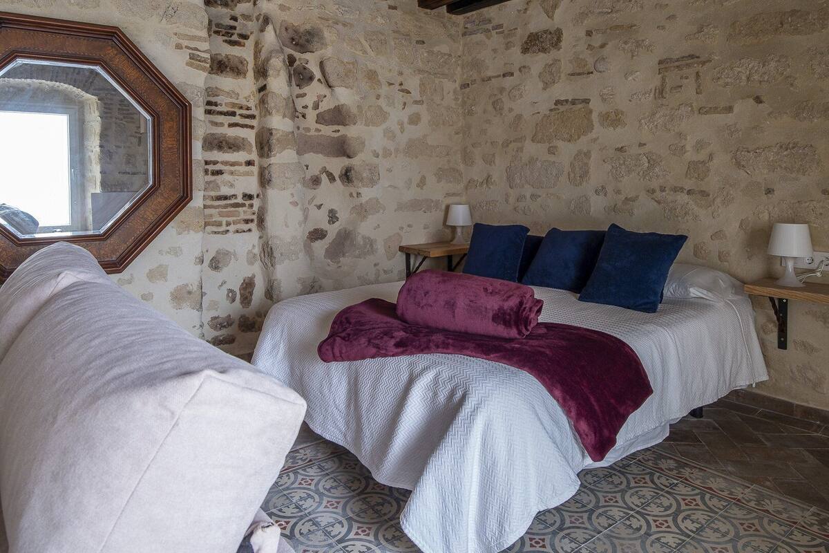 A room with a stone wall, a bed with a white and purple blanket, a wooden nightstand with a lamp, and a large mirror.