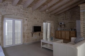 Un salon aux murs en pierre, avec un canapé blanc, une table basse en verre, un téléviseur et un coin cuisine doté de meubles en bois.