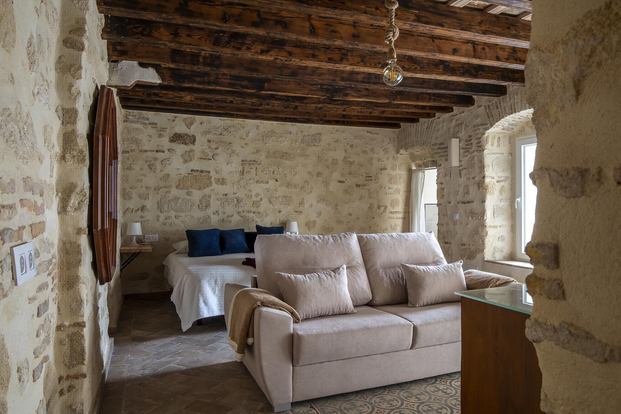 A rustic room with exposed wooden beams, a stone wall, a beige sofa, a bed with blue pillows, and a small table.