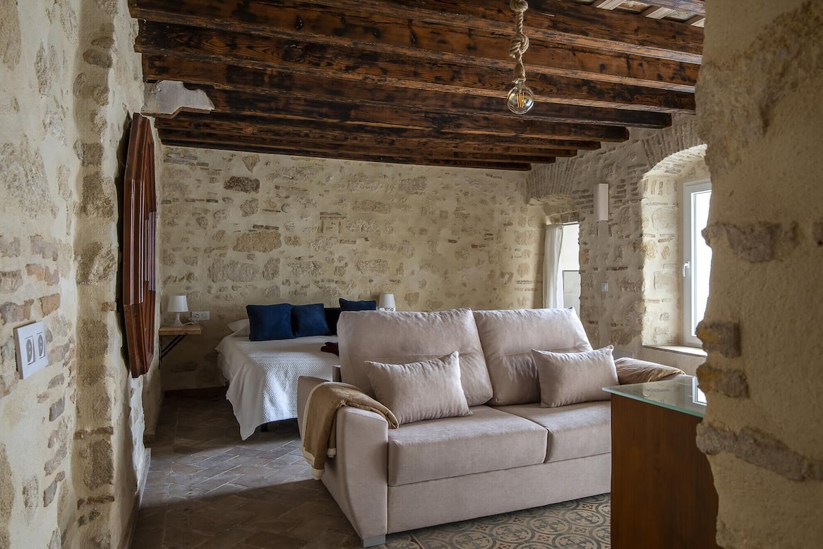 A rustic room with exposed wooden beams, a stone wall, a beige sofa, a bed with blue pillows, and a small table.