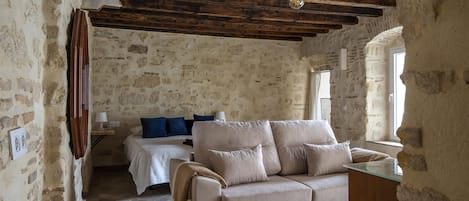 Une pièce rustique avec des poutres apparentes, un mur en pierre, un canapé beige, un lit avec des coussins bleus et une petite table.