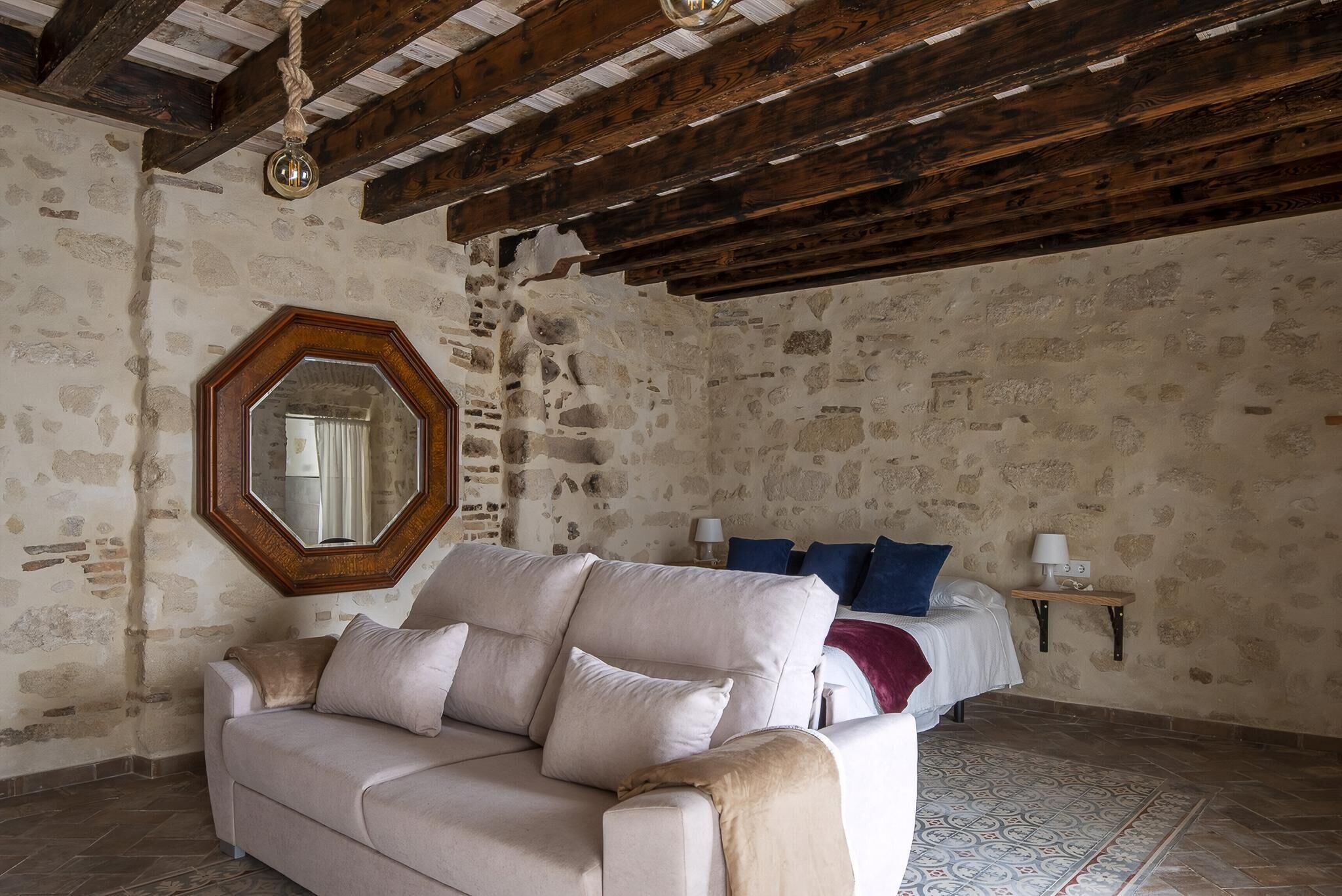 A room with a stone wall, wooden beams, a large mirror, a beige sofa, a bed with blue pillows, and a small table with a lamp.