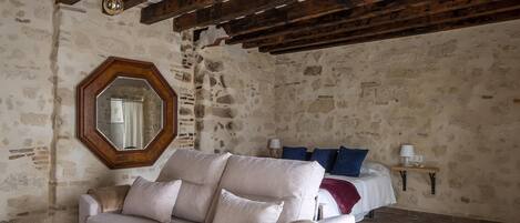 A room with a stone wall, wooden beams, a large mirror, a beige sofa, a bed with blue pillows, and a small table with a lamp.