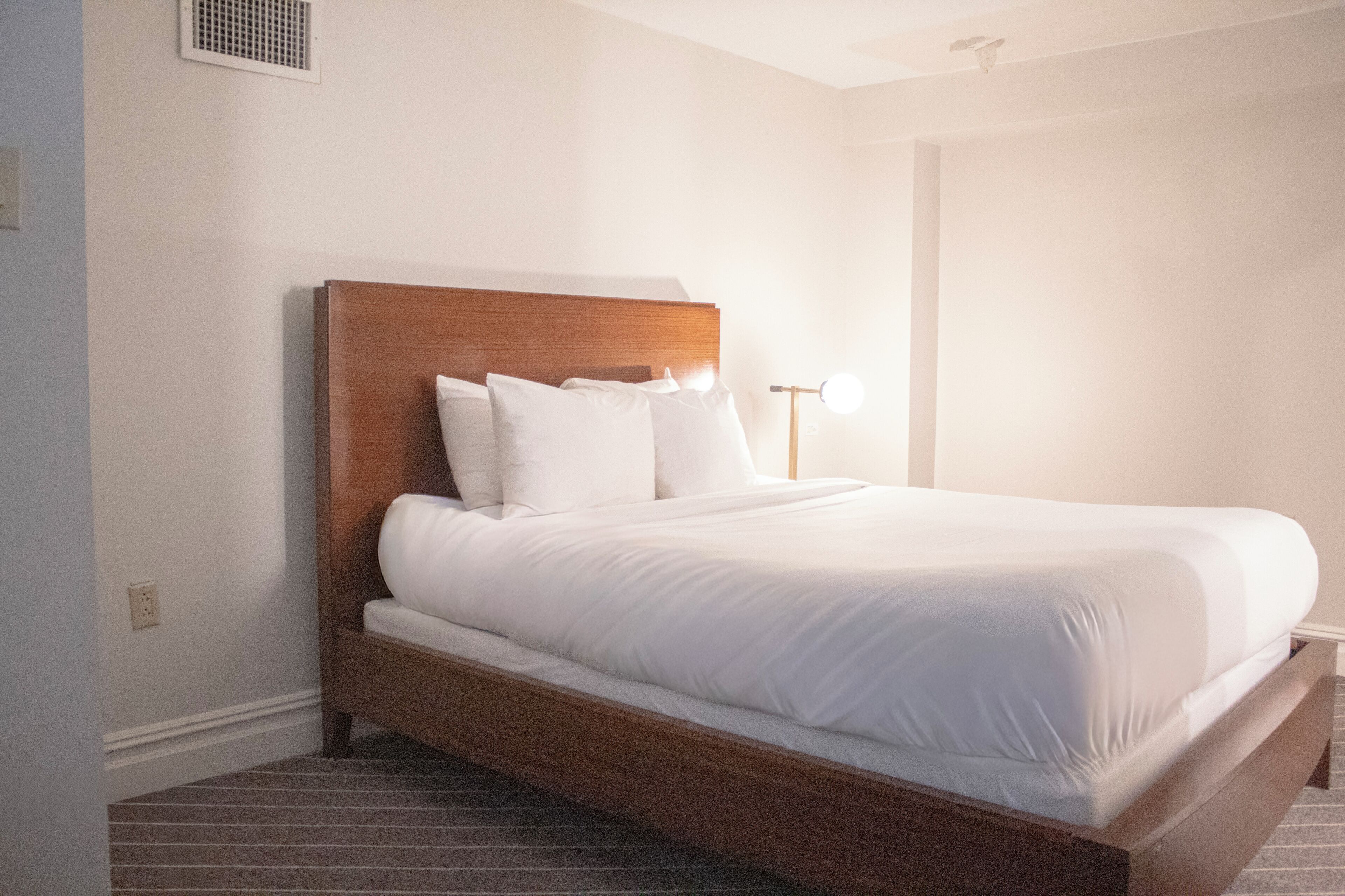A neatly made bed in a hotel room with a wooden headboard, a bedside lamp, and a visible power outlet on the wall.