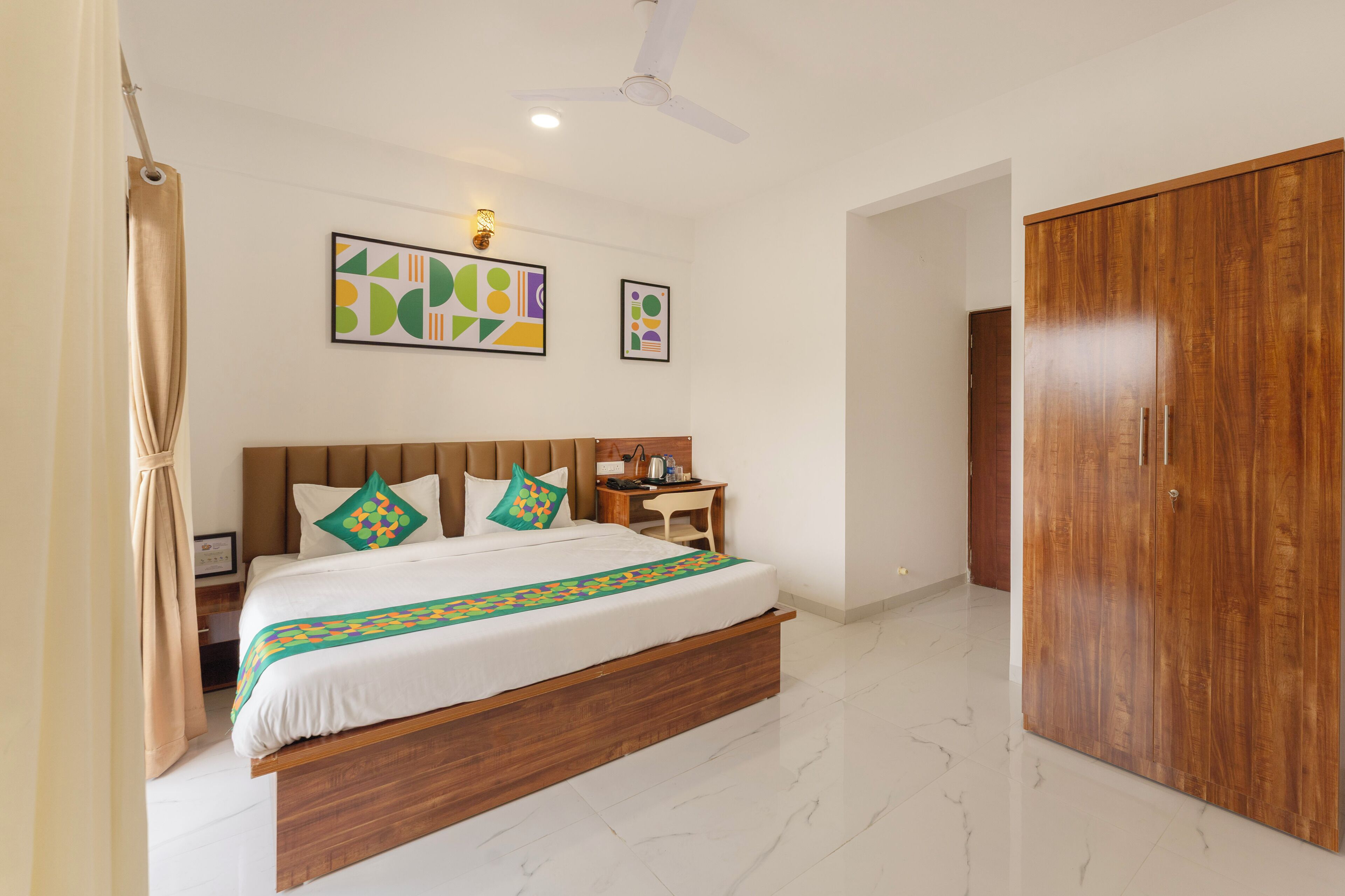 A bedroom with a bed, nightstand, desk, wardrobe, ceiling fan, artwork, and a bathroom door.