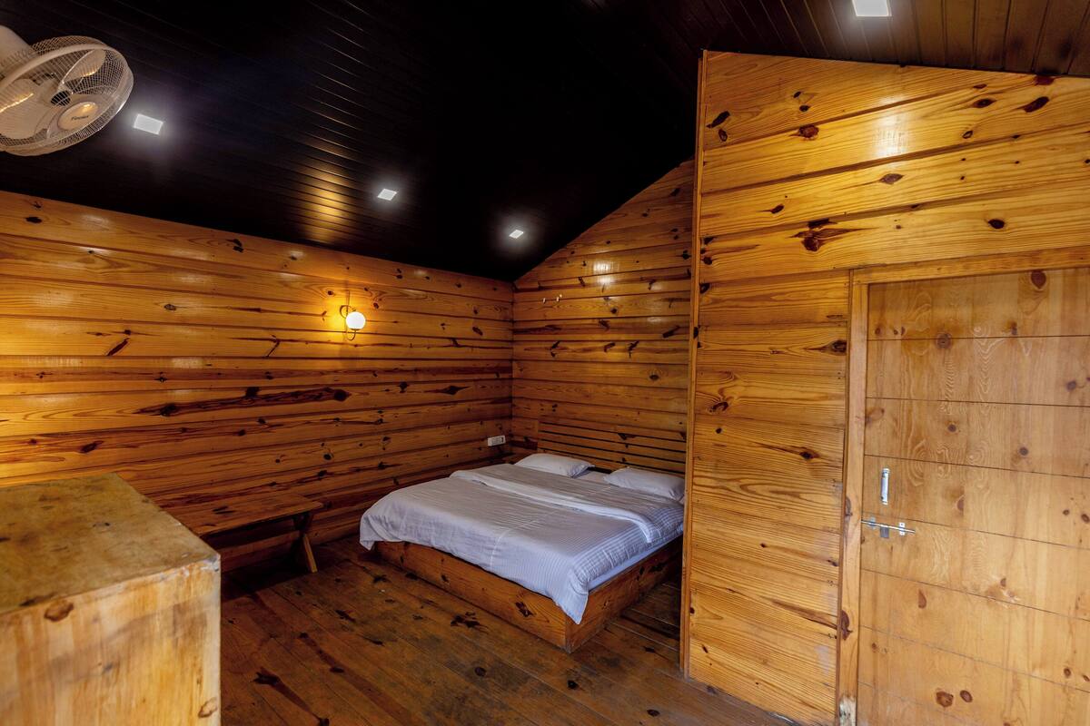 A wooden cabin room with a bed, a fan, and a small table.