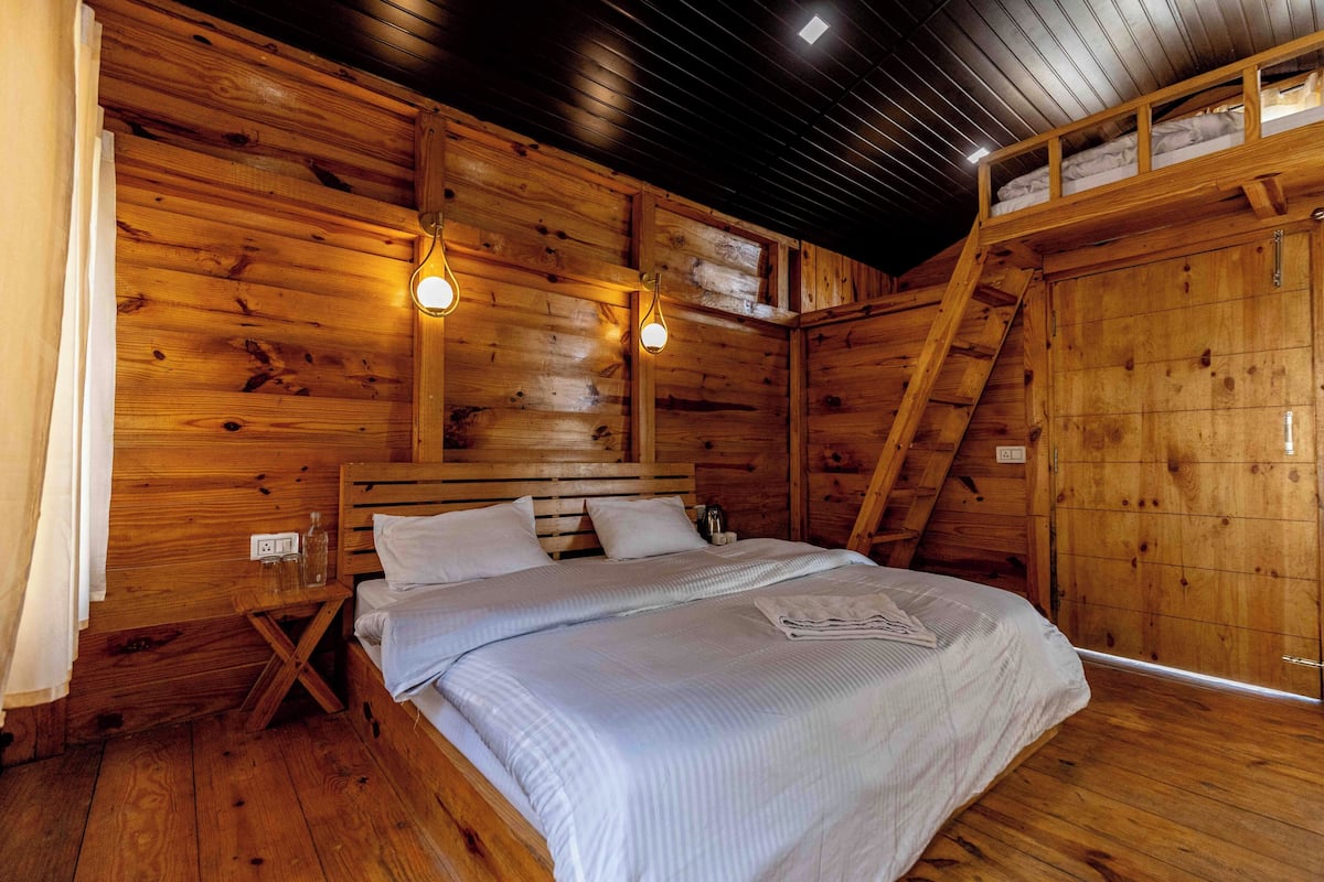 A wooden cabin bedroom with a bed, two wall-mounted lights, a small table, and a ladder.