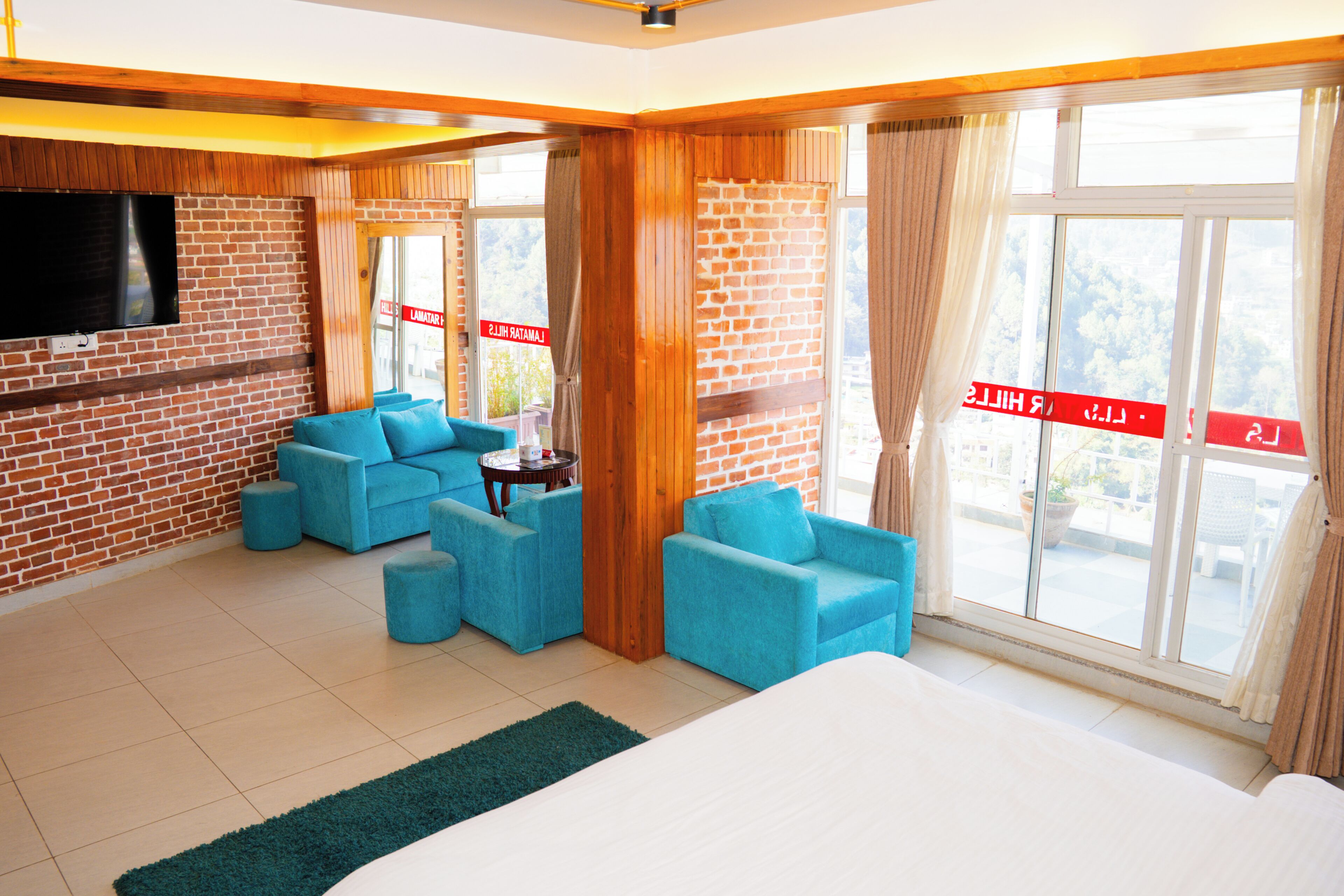 A hotel room with a brick wall, a flat-screen TV, a large bed, two blue armchairs, a small round table, and large windows with curtains.