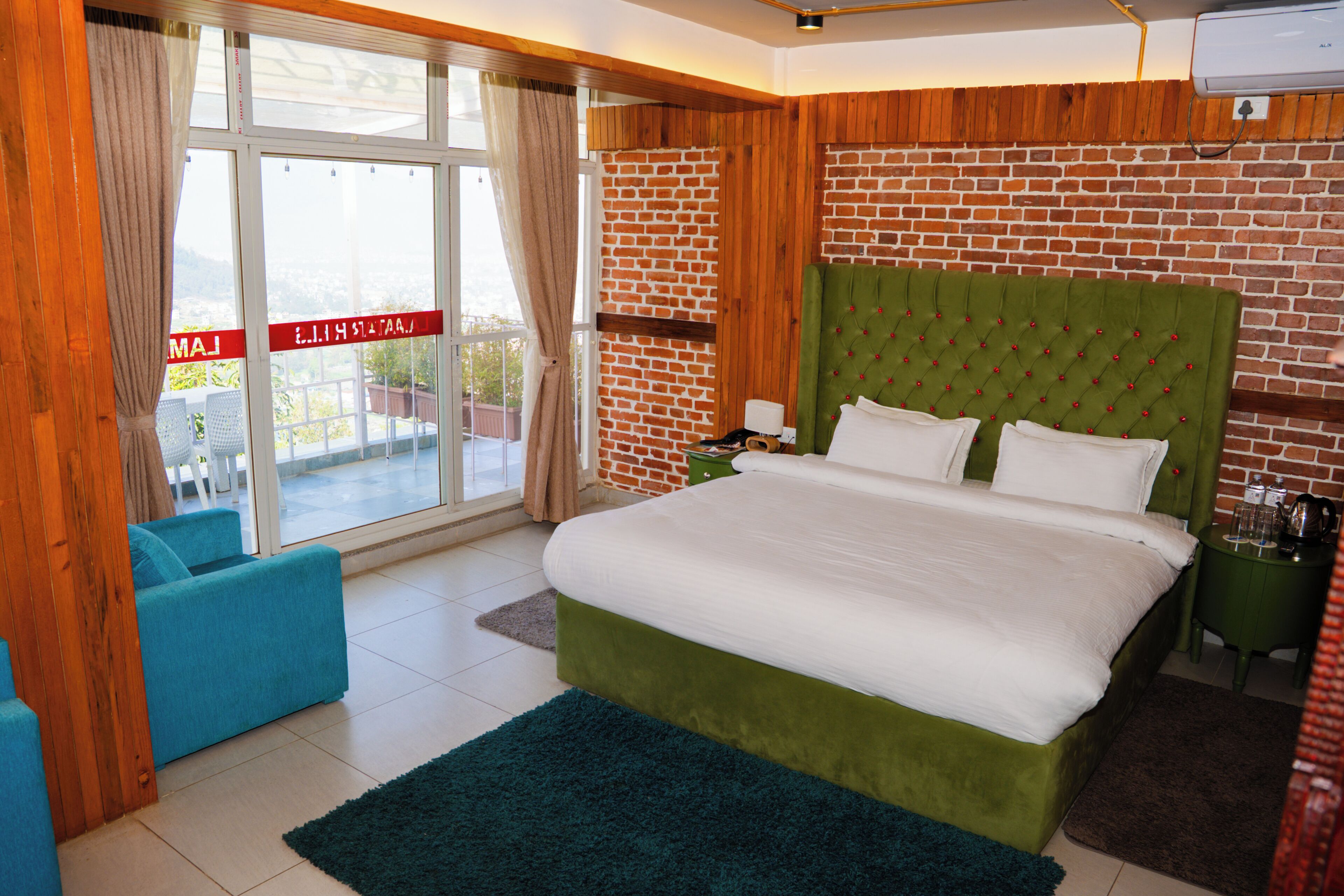A bedroom with a green upholstered headboard, a large bed, a blue armchair, a green nightstand, and a view through a glass door.