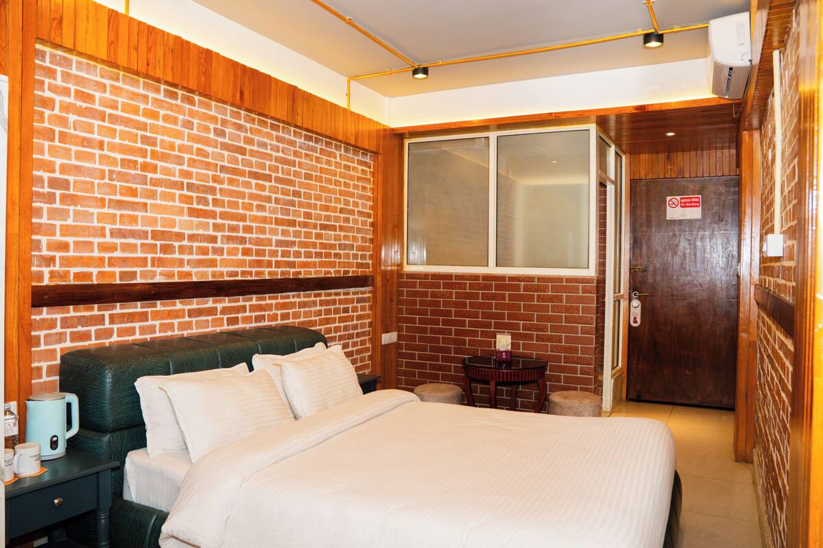 A hotel room with a brick wall, a bed with white bedding, a small table with a kettle, and a window.