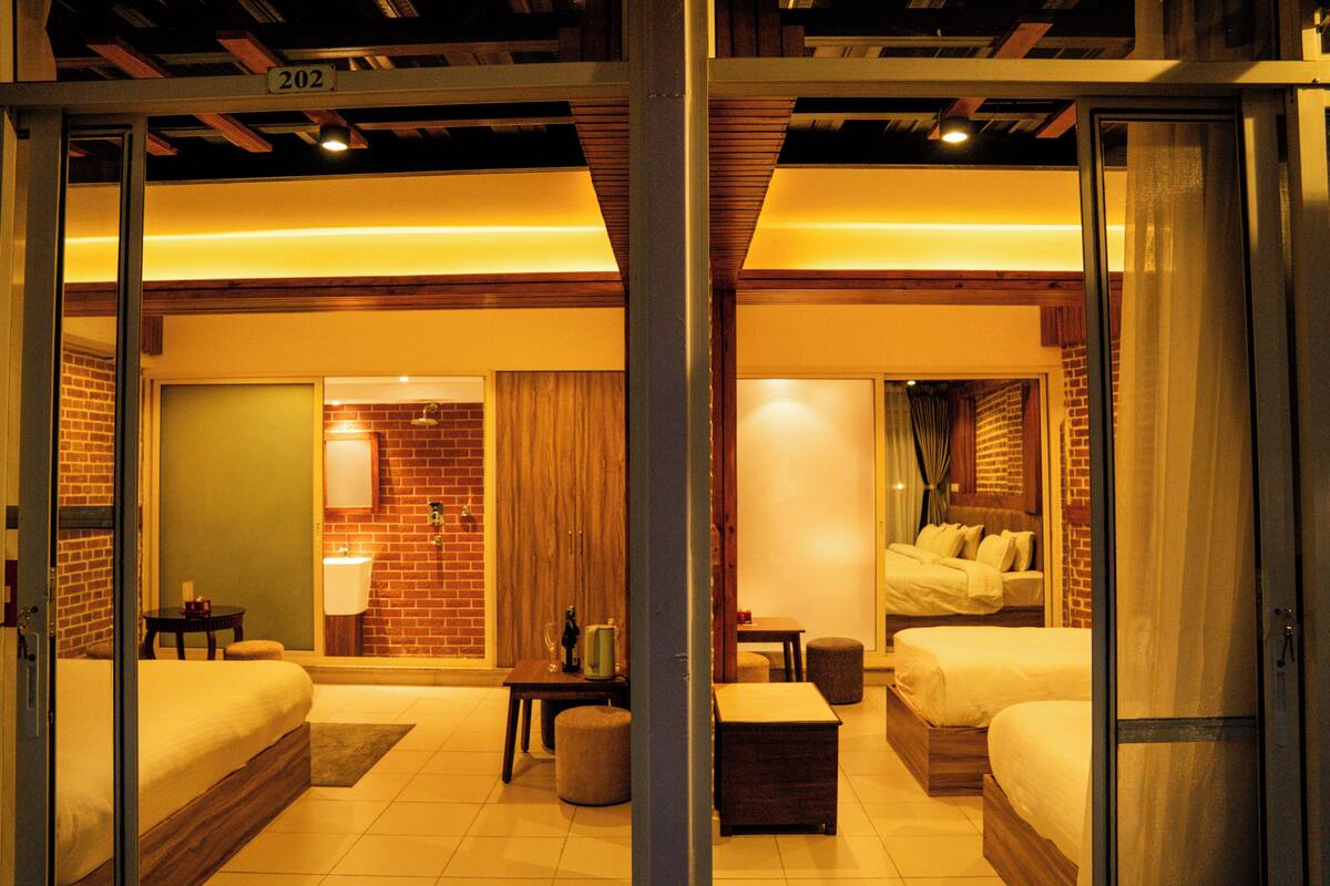 A hotel room with two beds, a small table, a chair, a sofa, and a bathroom with a mirror and sink.