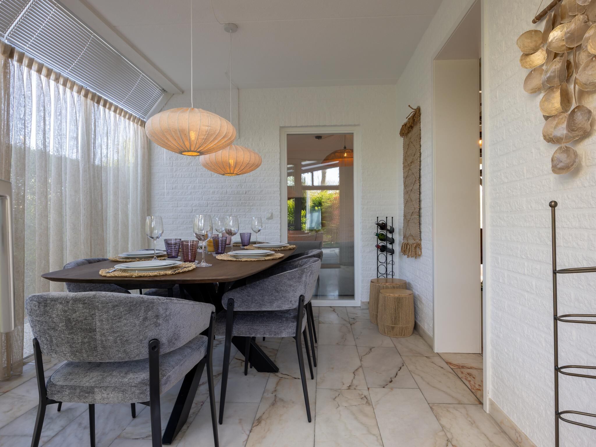 Een eetgedeelte met een marmeren vloer, grijze gestoffeerde stoelen, een houten tafel gedekt met glazen en borden, en hanglampen.