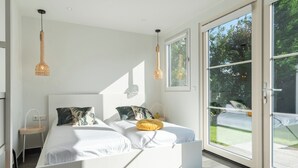 Ein modernes Schlafzimmer mit einem weißen Bett, zwei Pendelleuchten und einer großen Schiebeglasdoor, die zu einem Außenbereich führt.