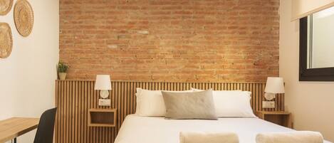 Une chambre d’hôtel avec un mur en briques, une tête de lit en bois, un lit avec du linge de maison blanc, deux tables de chevet avec des lampes, un bureau avec une chaise et une plante en pot.