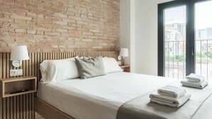 Ein Hotelzimmer mit Backsteinwand, einem Bett mit weißer Bettwäsche, zwei Nachttischlampen, einem Holznachttisch und Blick auf Gebäude durch ein großes Fenster.