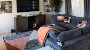 Ett vardagsrum med en platt-TV, en takfläkt, en soffa med kuddar, en växt och en matta.