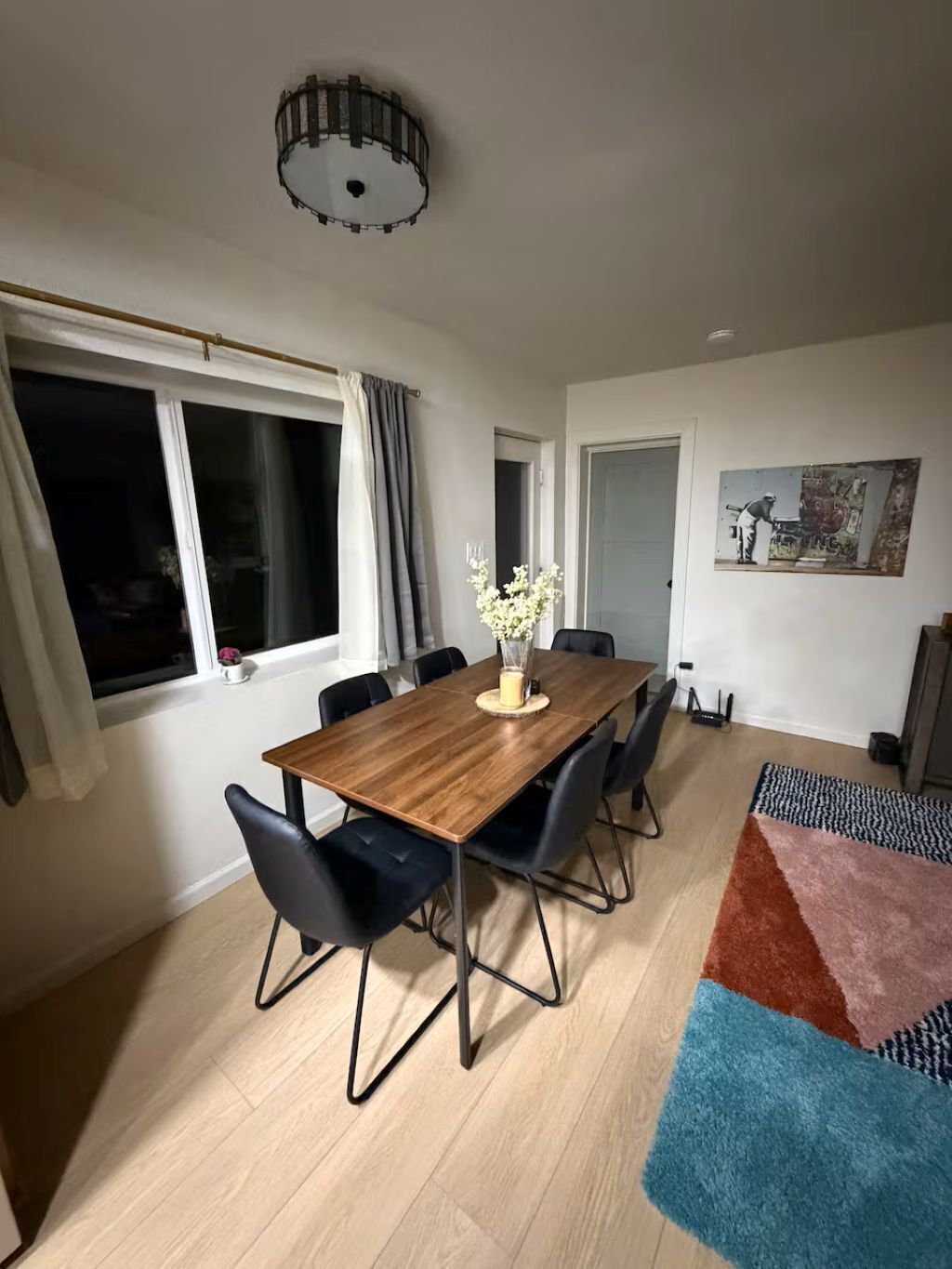 Een eetgedeelte met een houten tafel, zwarte stoelen, een vaas met bloemen, een plafondlamp en een schilderij aan de muur.