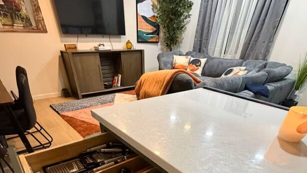 Ett kök med en inbyggd låda för köksredskap, en matplats med bord och stolar samt ett vardagsrum med en soffa och en tv.