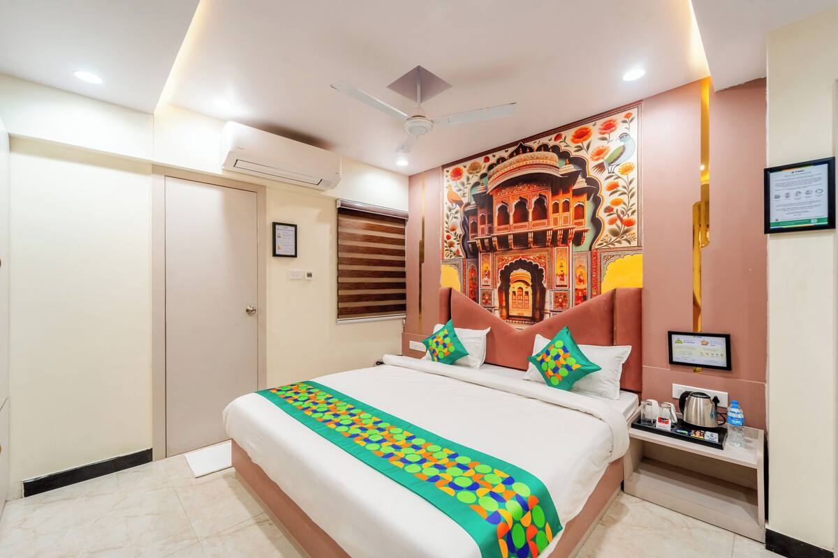 A hotel room with a bed, a ceiling fan, a wall tapestry, a bedside table with a kettle, and a framed certificate.