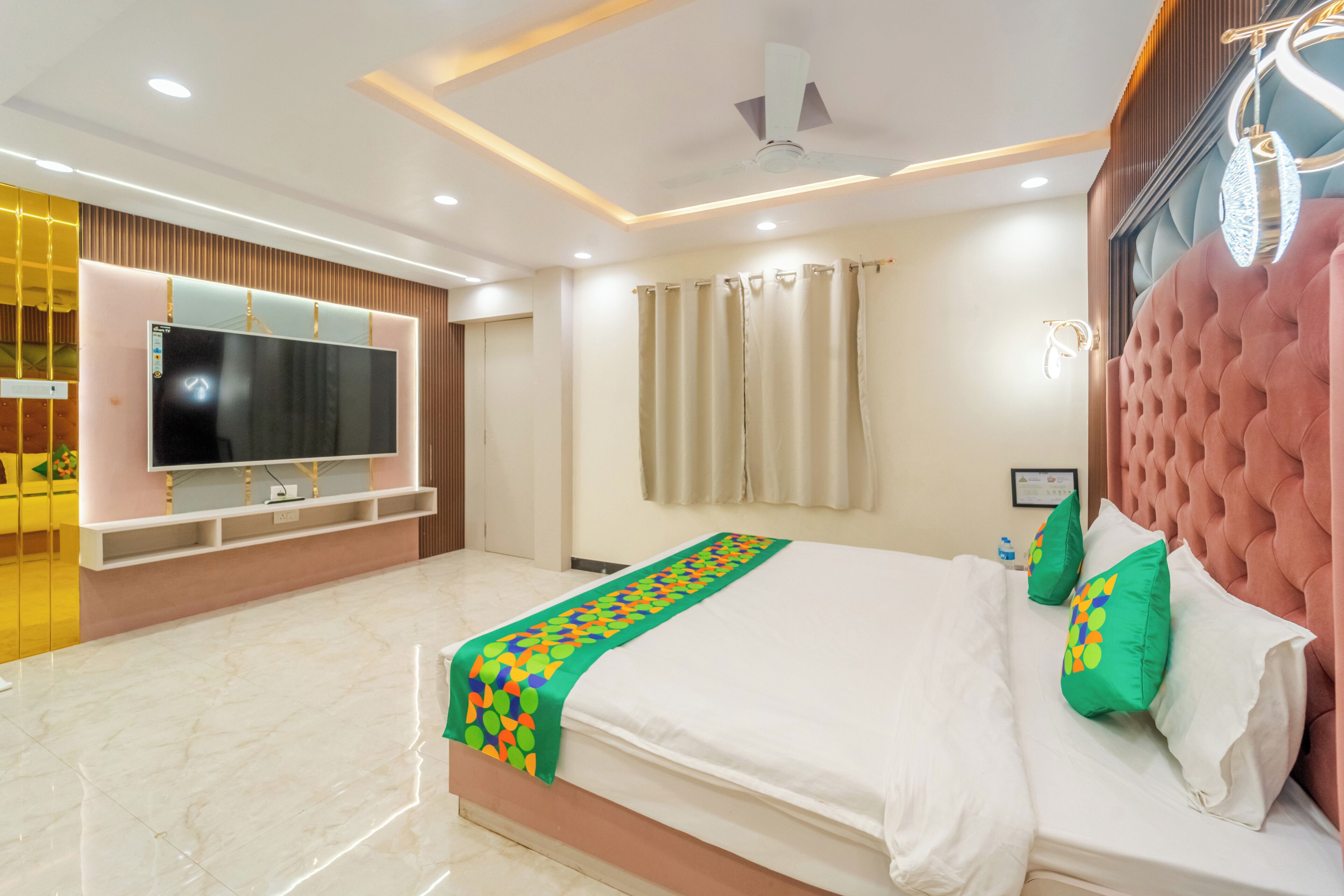 A modern hotel room with a large flat-screen TV, a bed with green and white bedding, a ceiling fan, and a large window with curtains.