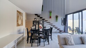 A modern dining area with a white table and black chairs, a sofa with cushions, a kitchen area with appliances, and a large window with a view. - Collection O near Sanur Beach formerly  (Denpasar)
