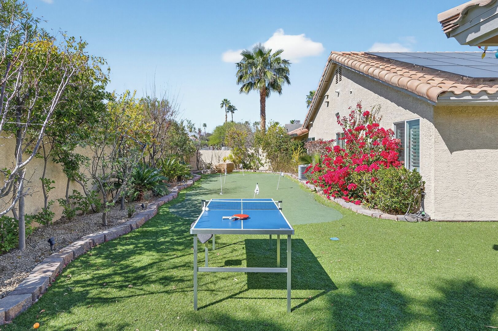 A ping pong table in a backyard with a house, palm trees, and flowering bushes.