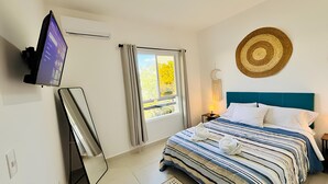 Quarto com cama, televisão na parede, janela com cortinas, objeto decorativo fixado na parede e mesa de cabeceira com abajur.