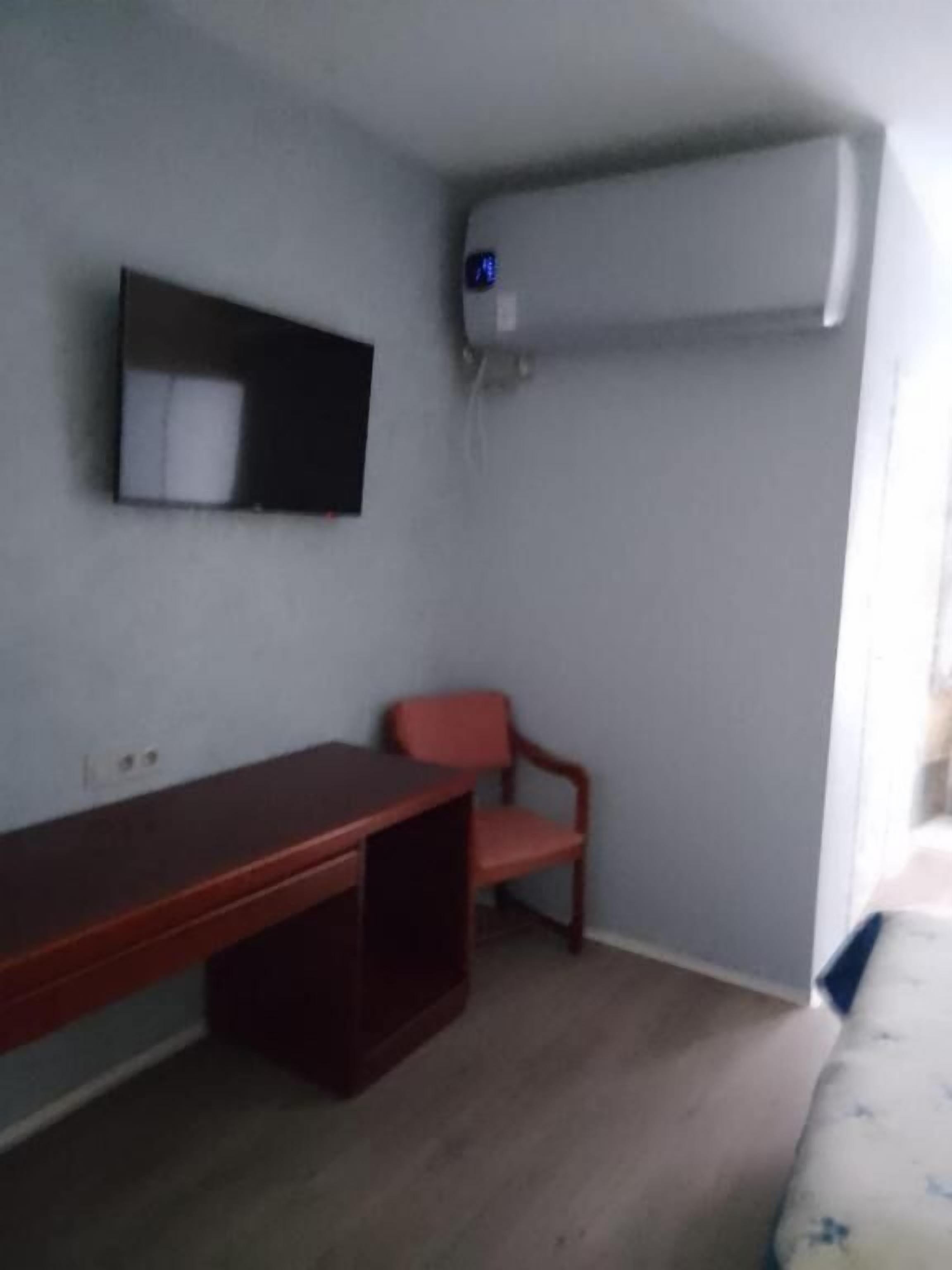 Une pièce avec un bureau en bois, une chaise rouge, un climatiseur fixé au mur et un lit recouvert d’une couvre-lit à motifs.