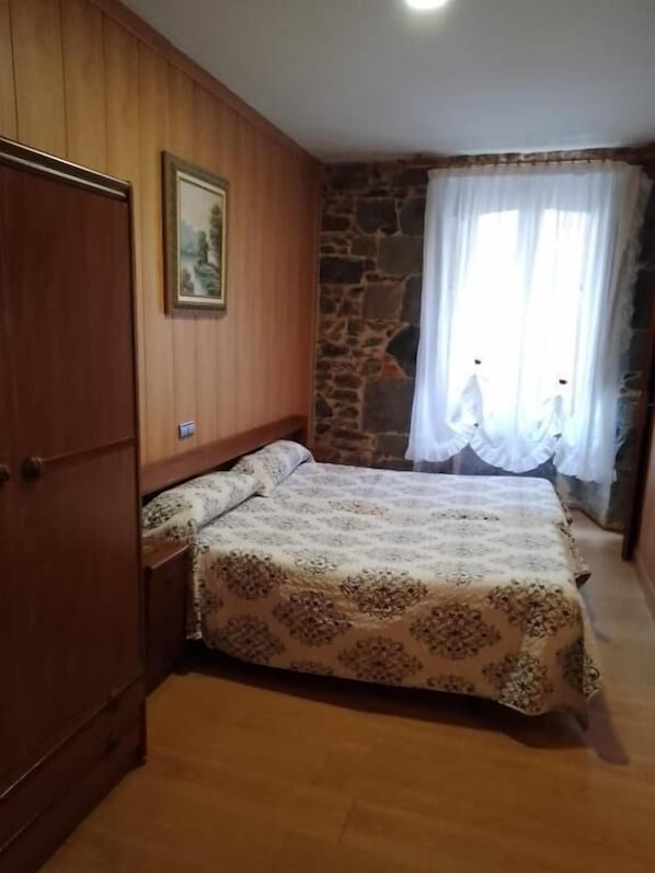 Un dormitorio con un armario de madera, una cama con colcha estampada, una pared de piedra y una ventana con cortinas blancas.