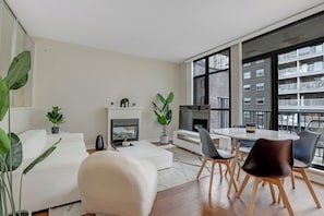 Ein modernes Wohnzimmer mit Kamin, einem Essbereich mit rundem Tisch und Stühlen sowie großen Fenstern mit Blick auf die Stadt.