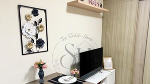 A modern living room with a flat-screen TV, a framed floral artwork, a small table with a vase of flowers, and a wall-mounted shelf with a sign. - High Floor Manila Bay View, Near MOA & Airport (Pasay City)