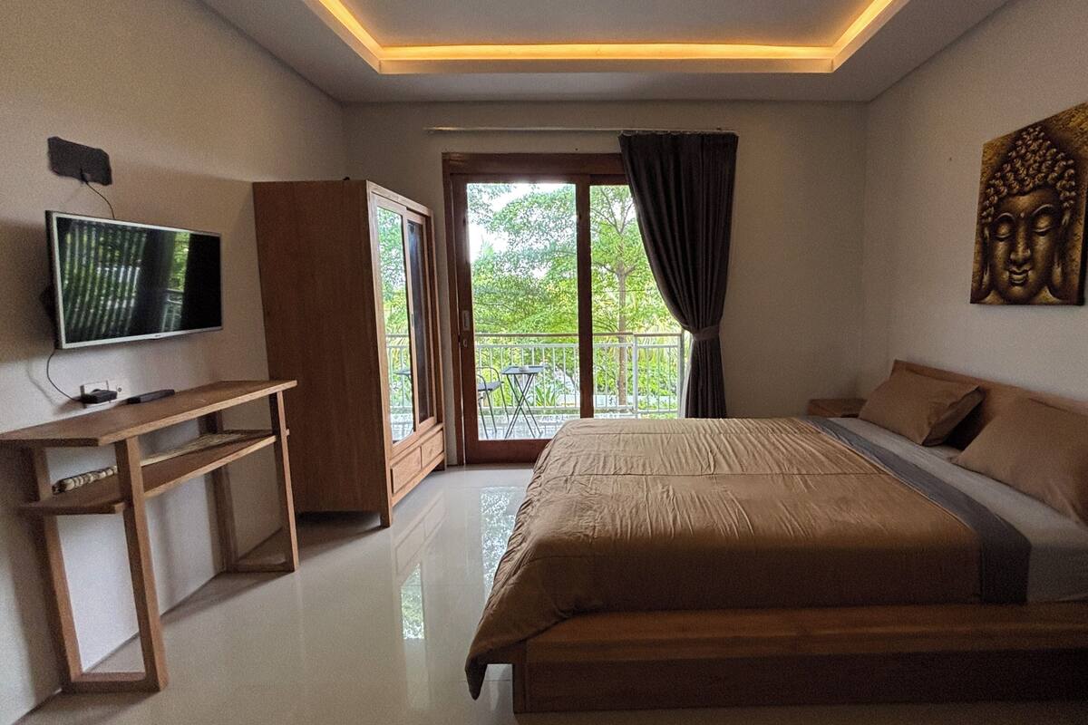 A bedroom with a bed, a TV, a desk, and a balcony with a view of greenery.