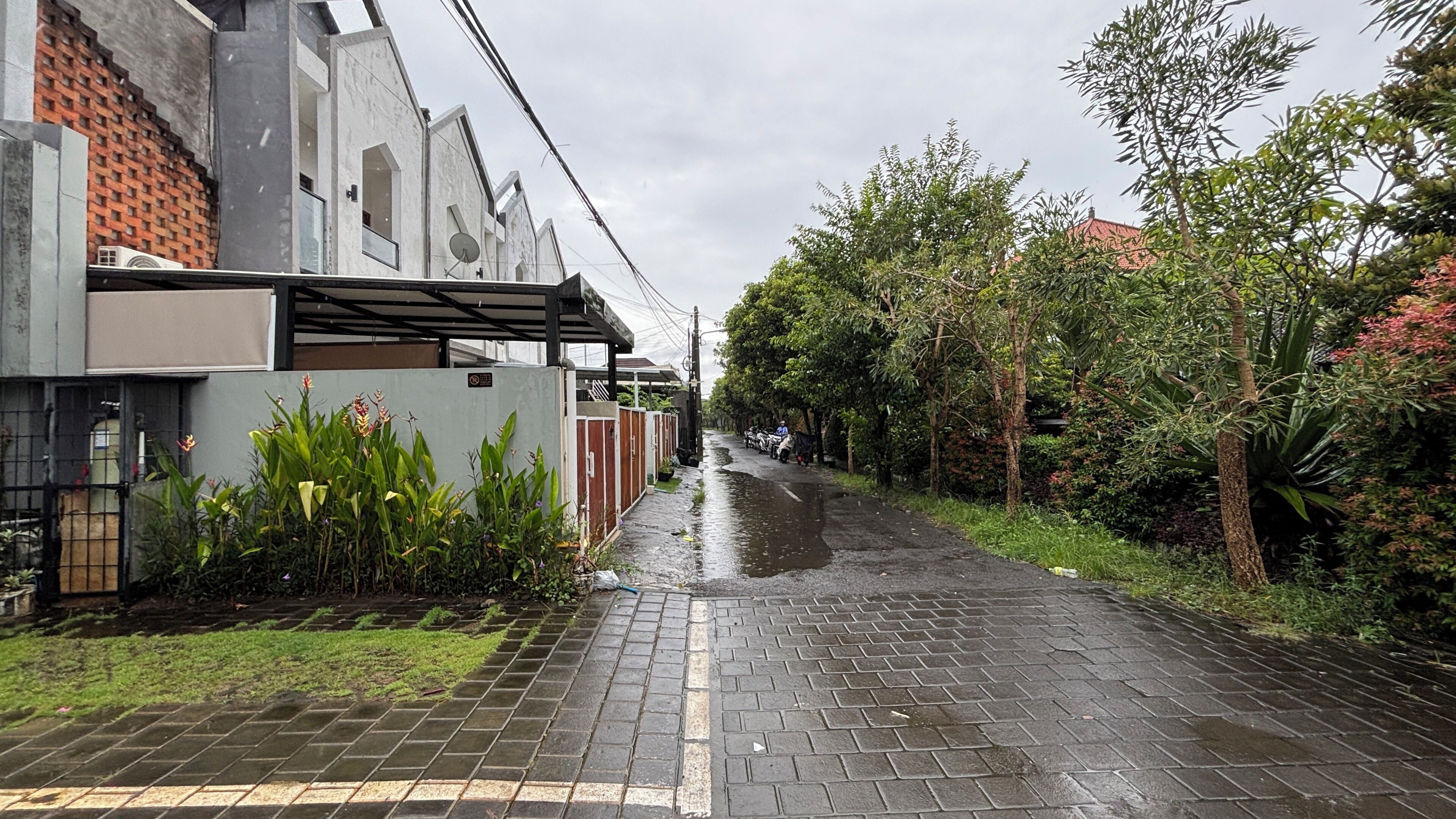 A residential street with a paved sidewalk, a brick wall, and a row of houses with satellite dishes.