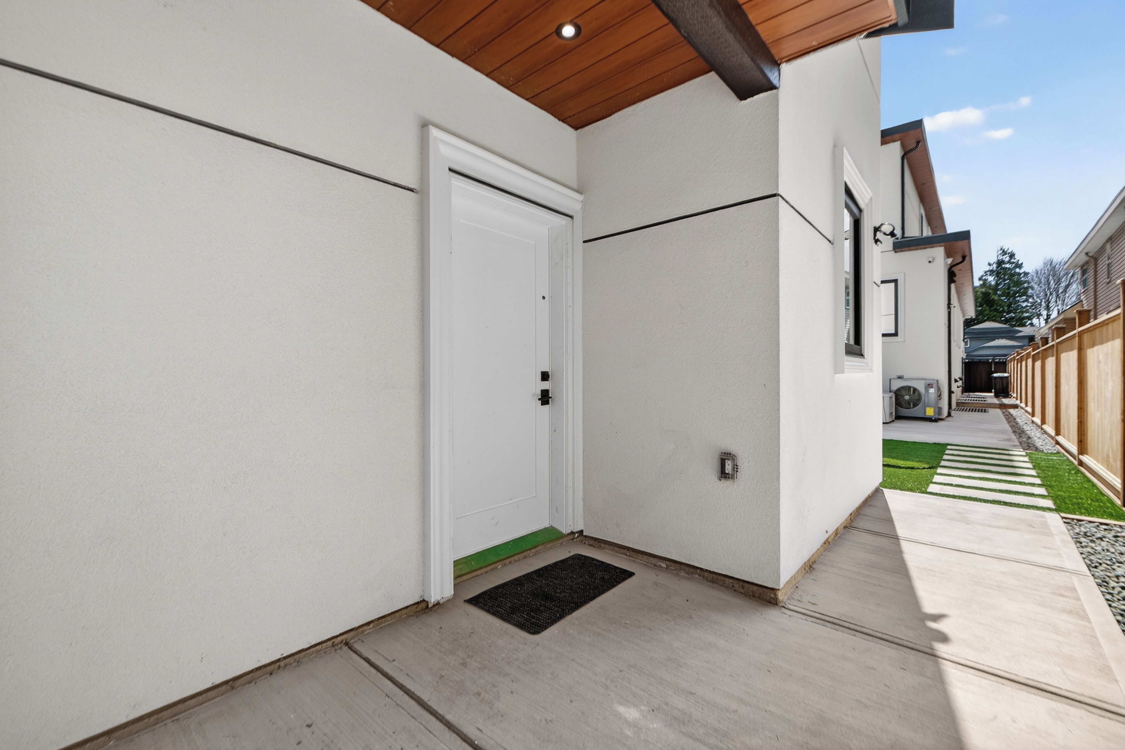 A clean, modern exterior with a white door, a wooden ceiling, and a concrete pathway leading to a green lawn.