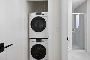 Ein Waschraum mit gestapelten Waschmaschinen und Trocknern, eine Dusche mit Glastüren und ein weiß gefliester Boden.
