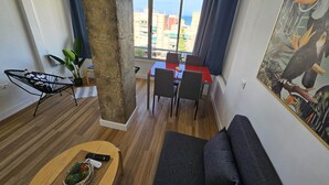 A modern living room with a wooden floor, a sofa, a dining table with chairs, a plant, and a large window with a view of buildings and the sea. - Apartment Torremolinos Near Bajondillo Beach (Torremolinos)