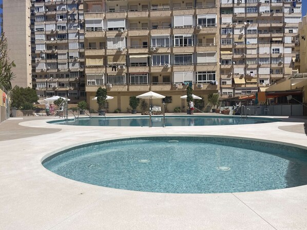 Ein Poolbereich mit einem runden Pool, umgeben von einem gepflasterten Weg, und ein mehrstöckiges Gebäude im Hintergrund.
