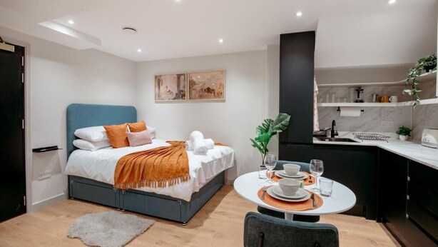 A modern hotel room with a bed, a dining table with chairs, a kitchen area, and artwork on the wall.