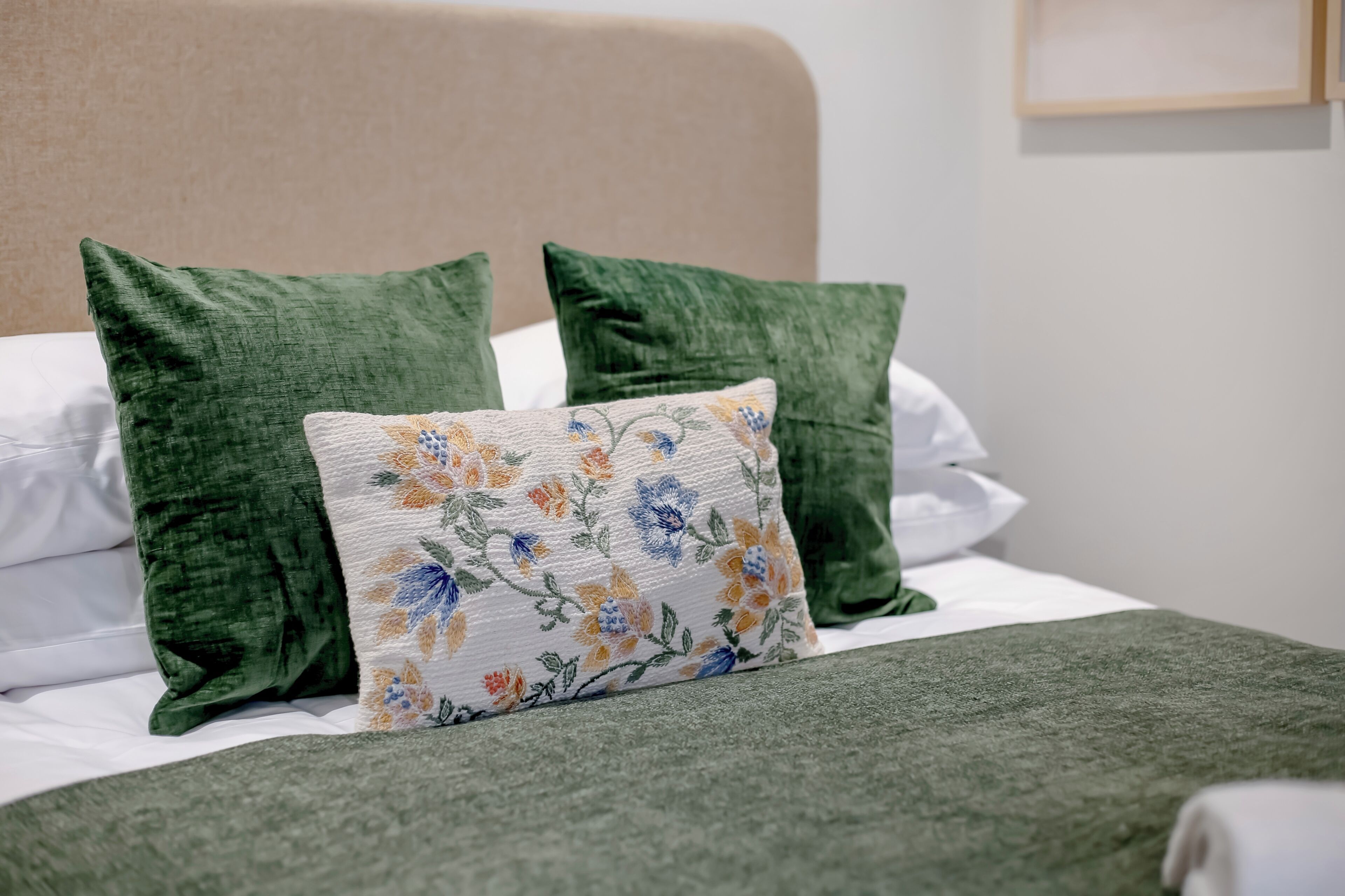 A bed with green pillows and a floral-patterned pillow, a beige headboard, and a white wall with a framed picture.