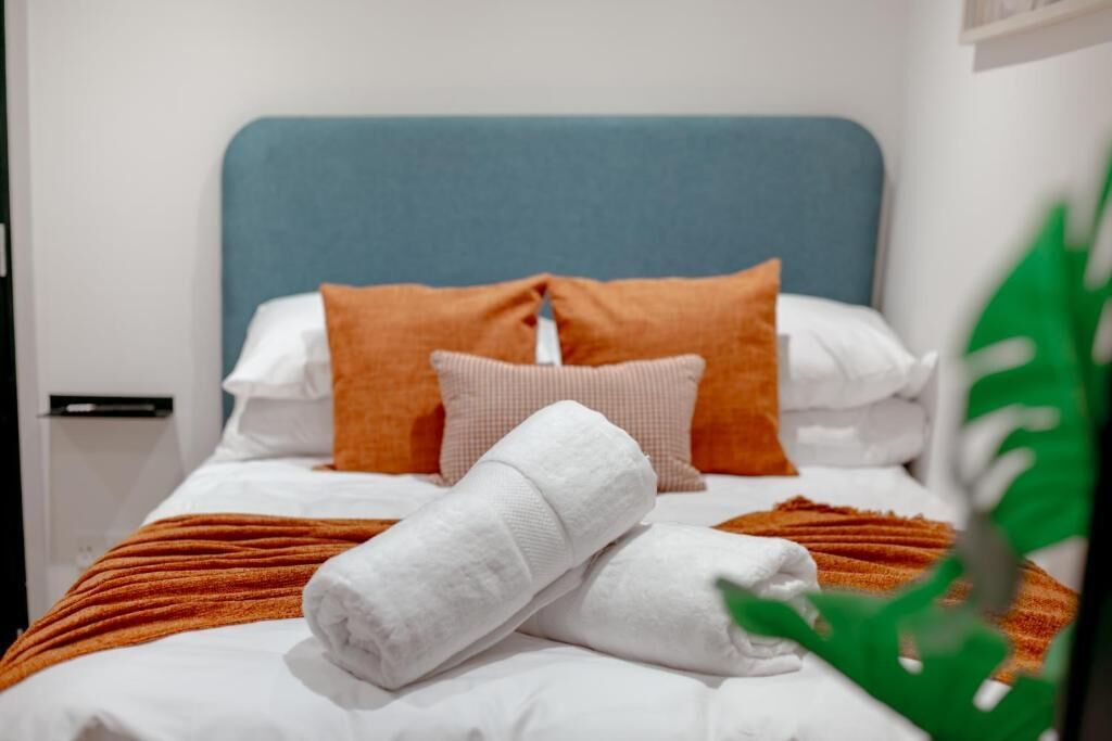 Un lit bien fait, avec une tête de lit bleue, des oreillers blancs et orange, et une serviette blanche.