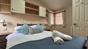 Ein Schlafzimmer mit Bett, Kissen, einem zusammengerollten Handtuch, einem Nachttisch, einer Lampe, einem Fenster mit Vorhängen und einer Tür.