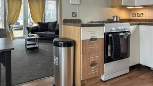 Een compacte keuken met een fornuis, oven en een vuilnisbak. In de aangrenzende woonkamer staat een kleine tafel en een bank.