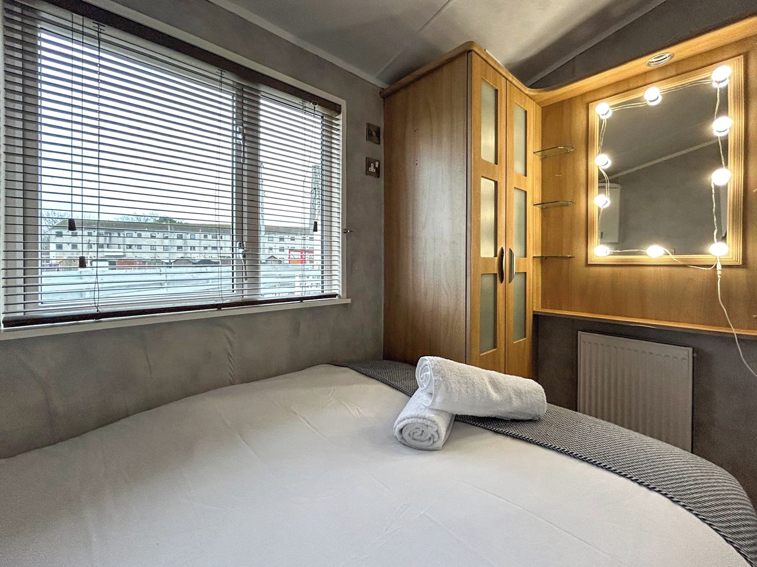 Een hotelkamer met een bed, een raam met luxaflex, een houten garderobe met spiegel en verlichting, en een radiator.